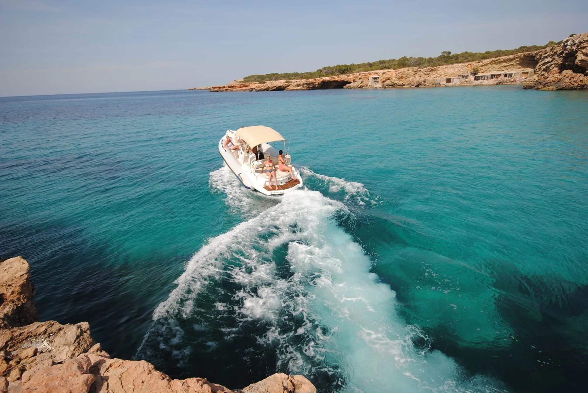 A charter boat cutting through clear turquoise water in a rocky Ibiza cala